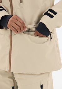 Beige ytterplagg med dragkedja framtill och en dragkedjeförsedd sidficka. Har svarta accenter på ärmarna och ett slätt, texturerat tyg.