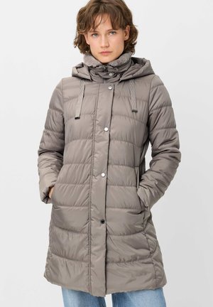 Young adult wearing a long gray padded winter coat with hood and hands in pockets standing against a plain background.