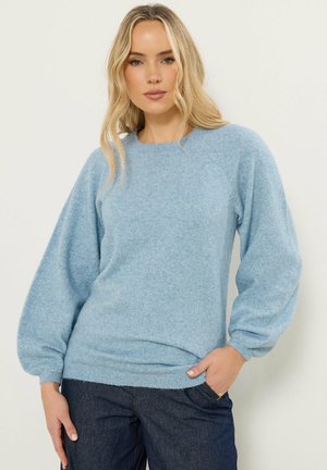 Donna bionda che indossa un maglione celeste a maniche lunghe con maniche a palloncino e jeans blu scuro, in piedi con una mano in tasca.