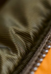 Teksturowana tkanina w zielono-brązowy wzór w kratę, obszyta brązowym ściegiem i z pomarańczowym akcentem. Miękka, lekko wyściełana w wyglądzie.