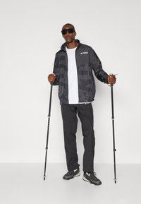 Schwarze leichte Jacke mit einem dezenten Muster, weißes T-Shirt, schwarze Hose und Wanderstöcke. Getragen mit Sonnenbrille und Wanderschuhen.
