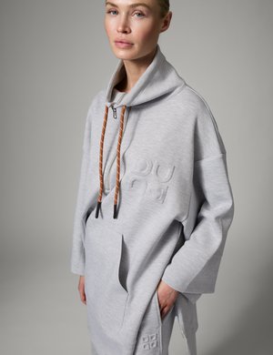 Kobieta w oversize jasnoszarej bluzie z kapturem z wysokim kołnierzem, brązowymi sznurkami, przednią kieszenią i wytłoczonym logo, stojąca na jednolitym tle.