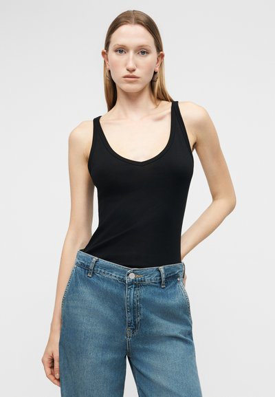 Γυναίκα που φοράει μαύρο αμάνικο τοπ και μπλε τζιν ψηλόμεσο, στέκεται με το ένα χέρι στην τσέπη μπροστά σε απλό φόντο.