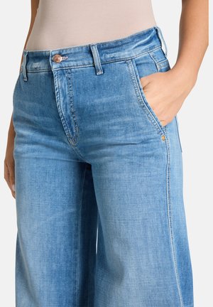 Frau trägt eine hochtaillierte hellblaue Jeans aus Denim mit einer Hand in der vorderen rechten Tasche, vor einem unifarbenen Hintergrund.