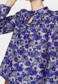 Blouse à manches longues en tissu à motifs avec des dessins floraux bleus et violets, dotée d'un col montant et d'une fermeture par bouton au niveau de l'encolure.