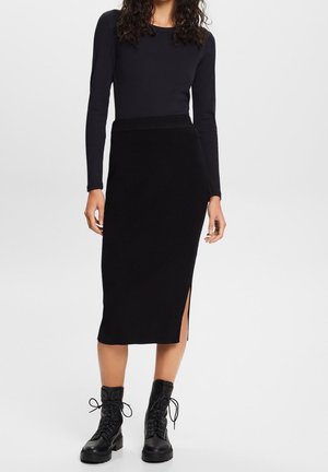 Zwarte top met lange mouwen, gecombineerd met een zwarte geribbelde midirok met zijslits, aangevuld met zwarte veterlaarzen tot aan de enkels.