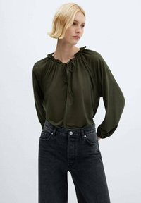 Bluză verde măslinie cu decolteu fluturat și detalii cu fundiță, mâneci lungi, asortată cu blugi negri din denim cu talie înaltă. Textură de material neted.