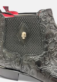 Bota negra con grabado floral texturizado, panel de malla y un detalle de calavera plateado. Presenta un diseño elegante y una superficie lisa.