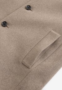 Cappotto di lana marrone con due bottoni neri, caratterizzato da una forma sartoriale e una tasca laterale. Il tessuto ha una finitura morbida e strutturata.