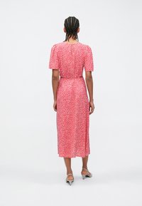 Κόκκινο μίντι φόρεμα με λευκές πουά βούλες, κοντά μανίκια με φουσκωτό στυλ και σφιγμένη μέση. Λείο ύφασμα με ελαφριά γυαλάδα, μήκος μέχρι το γόνατο.