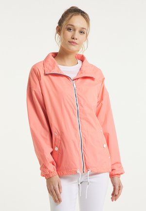 Jeune femme portant une veste zippée corail avec poches, poignets élastiques et cordons blancs, associée à un pantalon blanc, sur un fond clair.