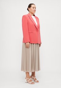 Koral blazer med en enkelt knapp, kombinert med en plissert beige skjørt og hvite stroppehæler. Glatt stoff, skreddersydd passform.