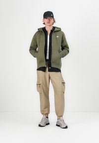Sweatshirt verde com capuz, camiseta preta, calças cargo beges e ténis cinzentos com detalhes pretos. Modelo está posicionado contra um fundo branco simples.