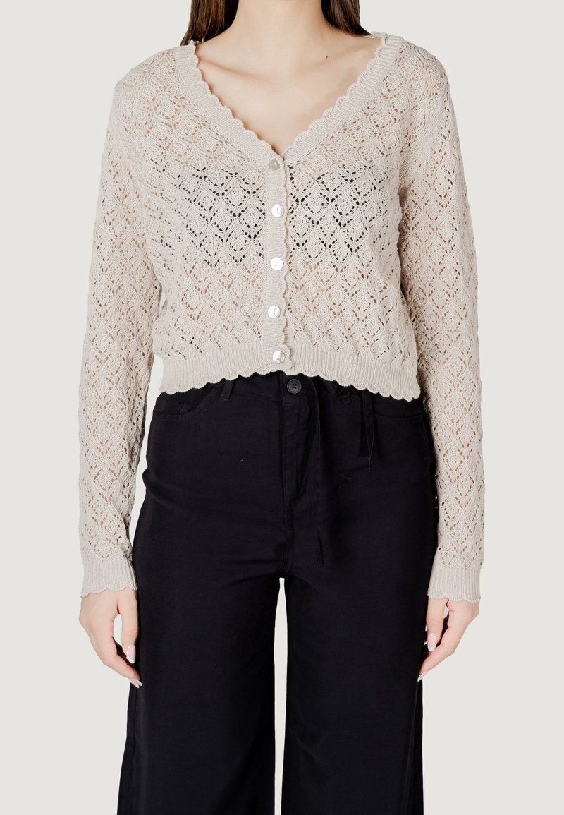 Cardigan en crochet beige boutonné avec bords festonnés porté avec un pantalon noir ample taille haute.