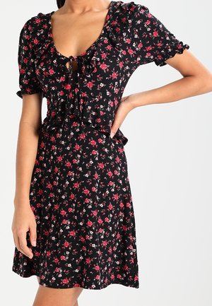 Robe fleurie noire avec manches courtes bouffantes, longueur mini, détail noué en forme de trou de serrure au niveau du col, ornée de motifs rouges et blancs sur un fond sombre.