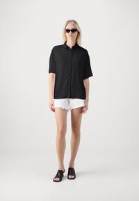 Svart kortermet skjorte med knappeopplegg og brystlomme, kombinert med hvite slitte shorts og svarte åpne mules.