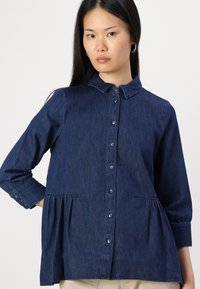 Denim shirt in dark blue, featuring a collared neckline, button front, long sleeves with button cuffs, and gathered waist detail.