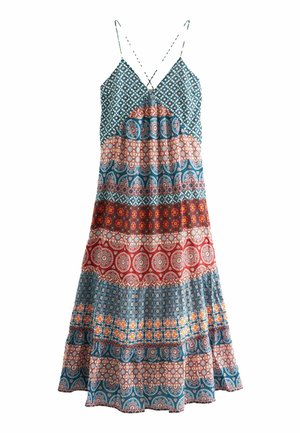 Mujer con un vestido maxi sin mangas de múltiples estampados, sosteniendo una bolsa tote negra con flecos, de pie en un patio de piedra cerca de una piscina.