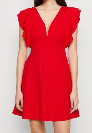 Kobieta w jasnoczerwonej sukience bez rękawów z falbanami na ramionach i głębokim dekoltem w kształcie litery V, stojąca na tle jasnego jednolitego tła.