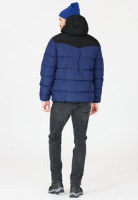 Blå puffjacka med svarta detaljer, featuring en huva och quiltad design. Bärs med mörkgrå jeans och svarta skor.