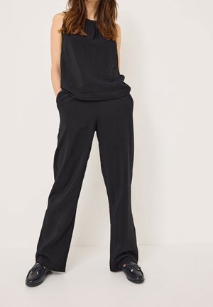 Vrouw die een mouwloze zwarte top en losse zwarte broek draagt, staand met handen in de zakken tegen een effen lichte achtergrond.