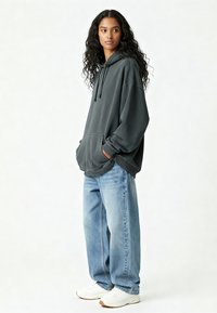 Donna con lunghi capelli ricci, che indossa un ampio hoodie grigio scuro, jeans larghi blu chiaro e sneakers bianche, in piedi con le mani in tasca.