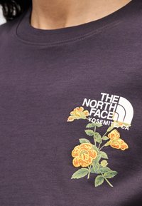 Μωβ βαμβακερό μπλουζάκι με λευκό λογότυπο "The North Face" και ζωντανή κίτρινη κεντητή ανθοδέσμη με πράσινα φύλλα, στρογγυλή λαιμόκοψη και κοντά μανίκια.