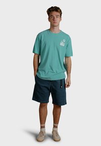 Camiseta de algodón color teal con mangas cortas y un gráfico blanco en el pecho izquierdo, combinada con pantalones cortos de mezclilla oscura y zapatillas color beige.