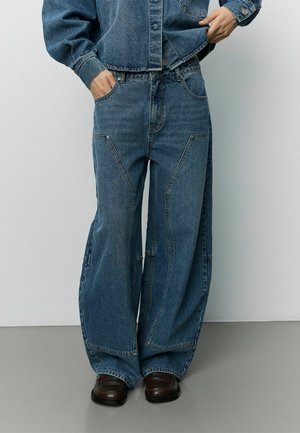 Personne portant un jean en denim bleu ample avec de grands panneaux cousus, une veste en denim assortie et des chaussures marron foncé, debout sur un sol gris.