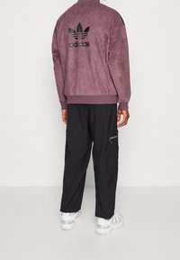 Lila mockaskinn Adidas-jacka med svart logotyp, kombinerad med svarta byxor med blixtlåsfickor och vita sneakers. Texturerad, sportig design.