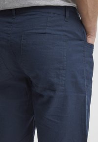 Tmavě modré kalhoty s jemnou texturou látky, vybavené zadní kapsou a volnějším střihem, upravené s standardními poutky na opasek a šicími detaily.