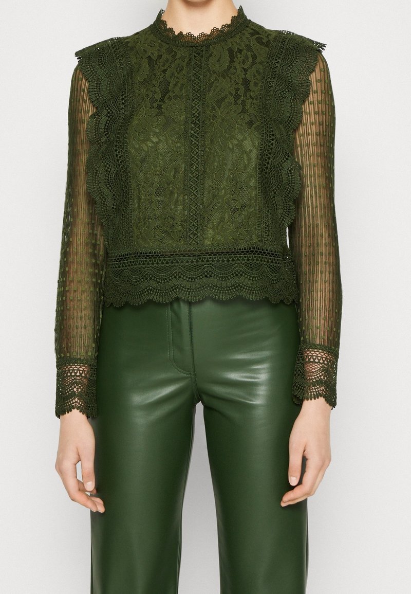 Top in pizzo verde scuro con maniche lunghe trasparenti, bordi a scallop e intricati motivi floreali. Abbinato a pantaloni in pelle verde coordinati.
