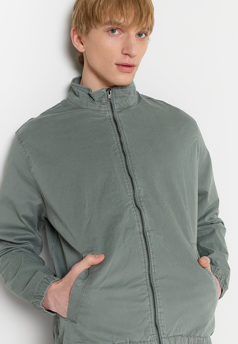 Hellgrüne Bomberjacke mit hohem Kragen, vorderem Reißverschluss und zwei seitlichen Taschen. Hergestellt aus einem weichen, glatten Stoff mit lässiger Passform.
