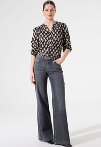 Schwarze und cremefarbene gemusterte Bluse mit Knopfverschluss, kombiniert mit hochgeschnittenen, lockeren, dunkelgrauen Jeans und schwarzen Schuhen.