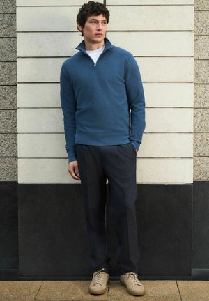 Man standing against a tiled wall wearing a blue quarter-zip sweater, dark pants, and beige sneakers, looking sideways with hands in pockets.