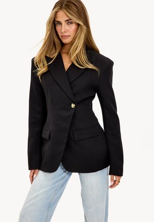 Blazer noir cintré avec un design croisé, col cranté, détails de boutons dorés et deux poches avant, associé à un jean bleu clair.