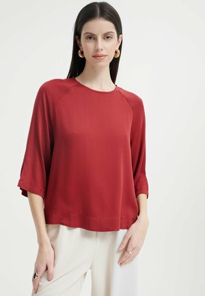 Maglietta rossa, leggera, con vestibilità comoda, maniche a 3/4, scollatura rotonda e una leggera lucentezza. Indossata sopra pantaloni bianchi ampi.
