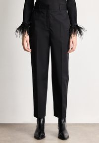 Persona valkā melnas piegrieztas bikses, melnus smailu purngalu potīšu zābakus un melnu ribotu džemperi ar spalvām rotētām aprocēm.