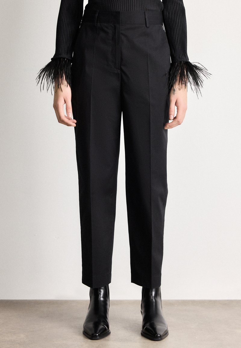 Persona valkā melnas piegrieztas bikses, melnus smailu purngalu potīšu zābakus un melnu ribotu džemperi ar spalvām rotētām aprocēm.