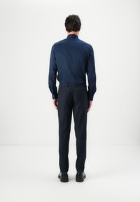 Cămașă albastru marin cu nasturi și mâneci lungi, purtată cu pantaloni închiși la culoare. Material neted cu cusături minime vizibile și două buzunare la spate.