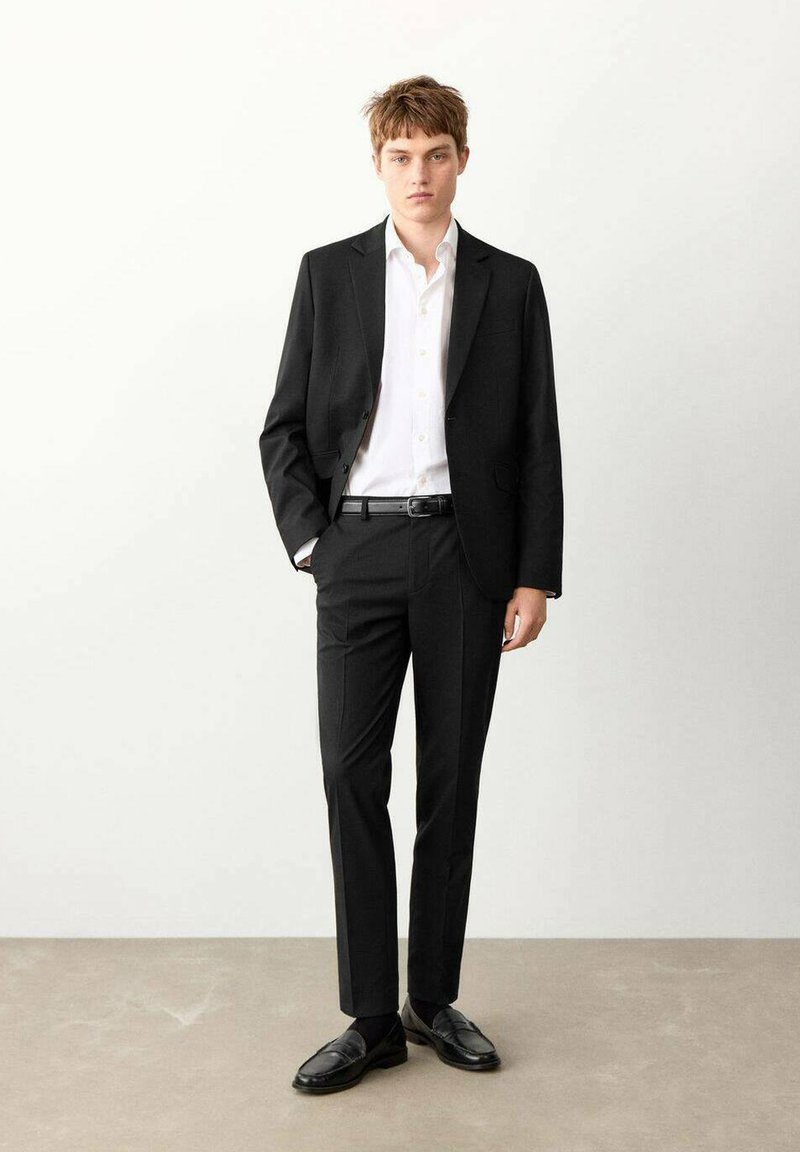 Costume noir avec une veste cintrée et un pantalon slim, associé à une chemise blanche à boutons et des chaussures en cuir noir. Texture du tissu lisse.