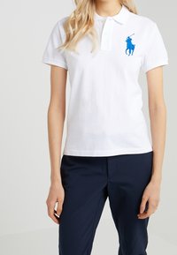 Vit pikétröja i bomull med två-knapps krage. Har en blå broderad häst och ryttare-logga på vänster bröst.