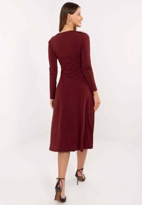 Bordowa sukienka midi z długimi rękawami, dopasowanym gorsetem, marszczonym tyłem i gładką fakturą. W zestawie z czarnymi butami na obcasie.