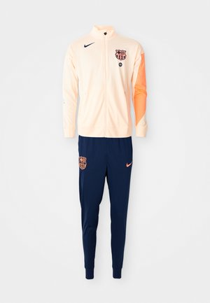 Light peach zip-up jacket with navy blue pants. Features logo accents, contrasting sleeve detail, and fitted cuffs. Sports material.