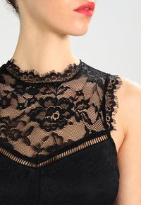 Haut en dentelle noire avec un design à col haut, orné de motifs floraux, de bords effilochés et d'une texture transparente, complété par une base noire unie.