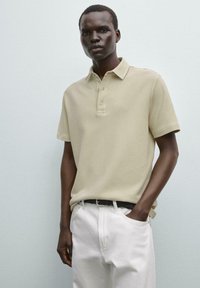 Beige polo tričko s ribkovanou texturou, límcem na tři knoflíky a krátkými rukávy, spárované s bílými kalhotami a černým opaskem.