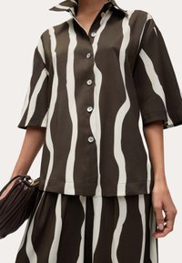 Camicia a maniche corte a righe marroni e bianche con colletto alto, tessuto morbido e bottoni nascosti. Abbinata a una gonna coordinata.