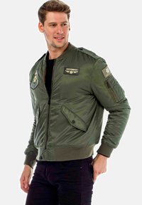 Olijf groene bomberjack met een rits aan de voorkant, gebreide manchetten en zakken. Bevat patches op de borst en mouw, glanzend nylon materiaal.