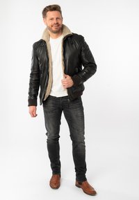 Chaqueta de cuero negra con forro de felpa beige, puños acanalados y dos bolsillos frontales, llevada sobre una camisa blanca y combinada con jeans oscuros y botas marrones.