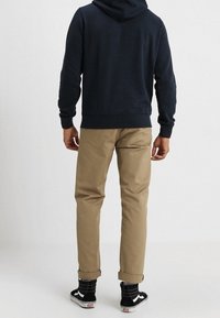 Person iført en navy hættetrøje, beige bukser med opkrogede kanter, sorte og hvide sneakers, stående med ryggen til mod en ensartet lys baggrund.